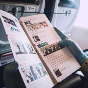 A passenger's hands holding an open, multi-page instructional brochure on an airplane, featuring clean layout design with illustrations, icons, and structured text by AUZ Studio.