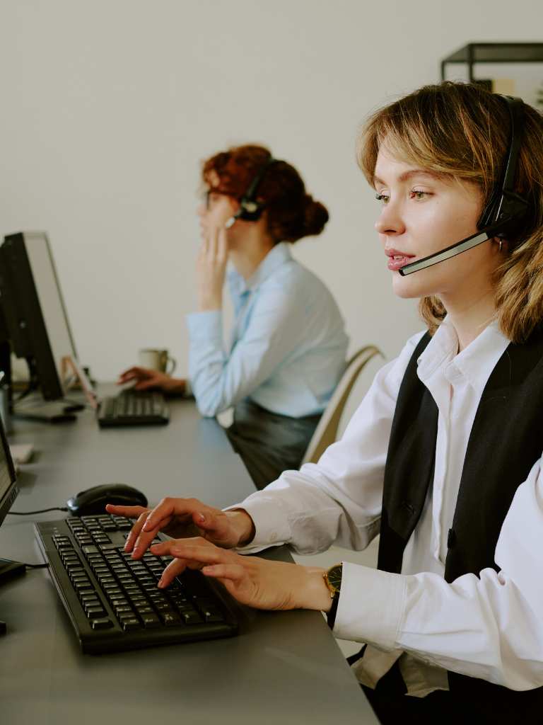 A professional woman in a white shirt and black vest wearing a headset and typing at a computer, providing dedicated customer support in a modern AUZ Studio office setting.