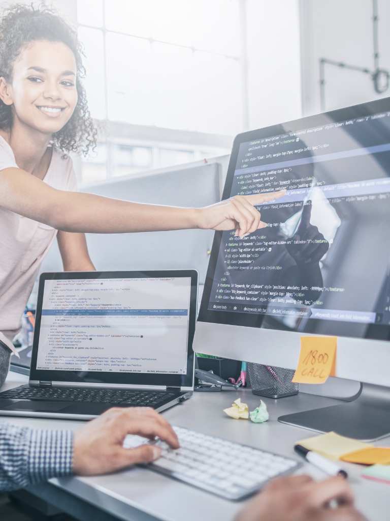 A female developer smiling while pointing at complex lines of code on a large monitor, collaborating with a male colleague in a bright, modern office space to build a website for AUZ Studio.